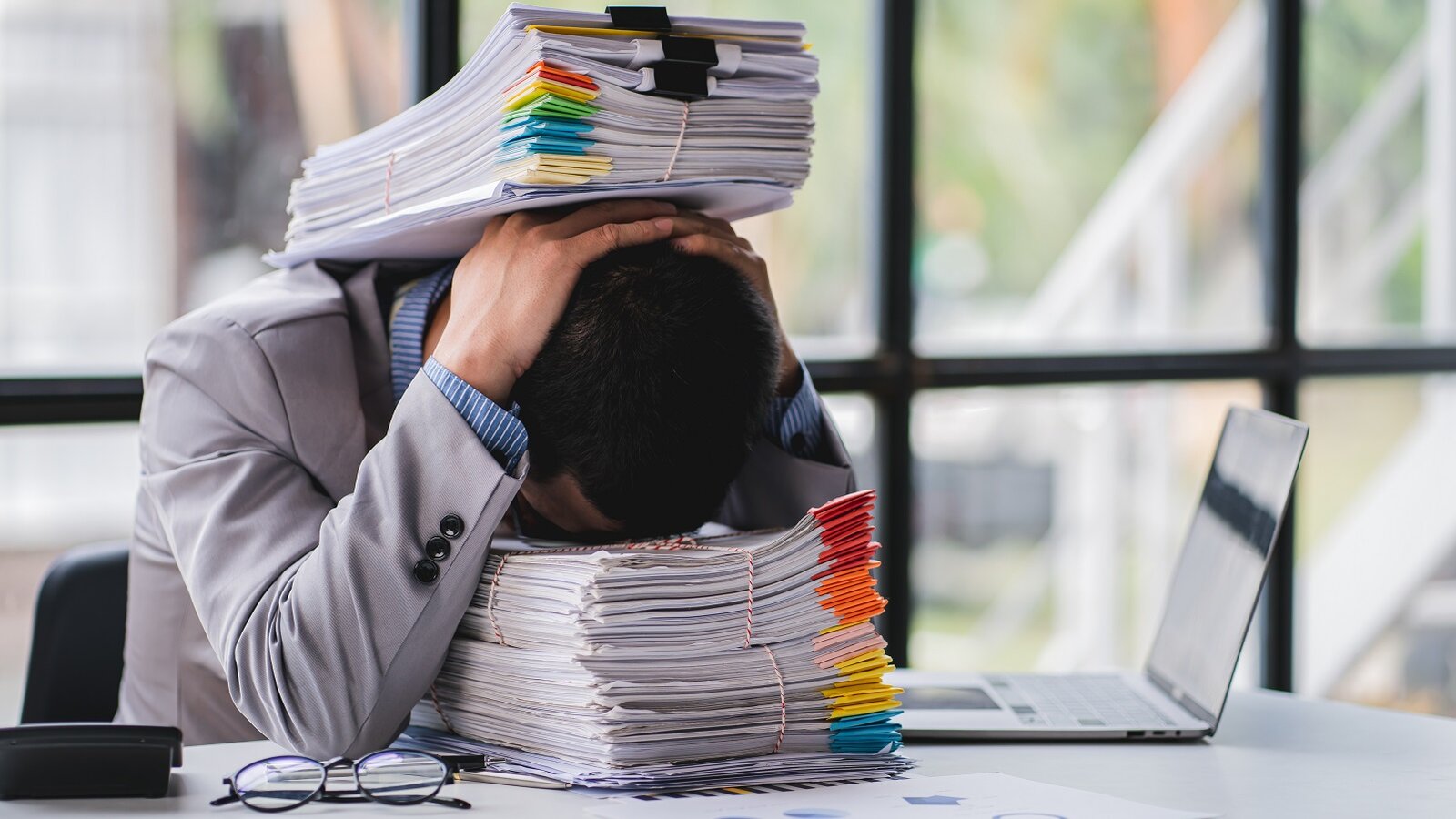 Mann lehnt den Kopf auf Formulare, weiterer Formularstapel liegt auf ihm